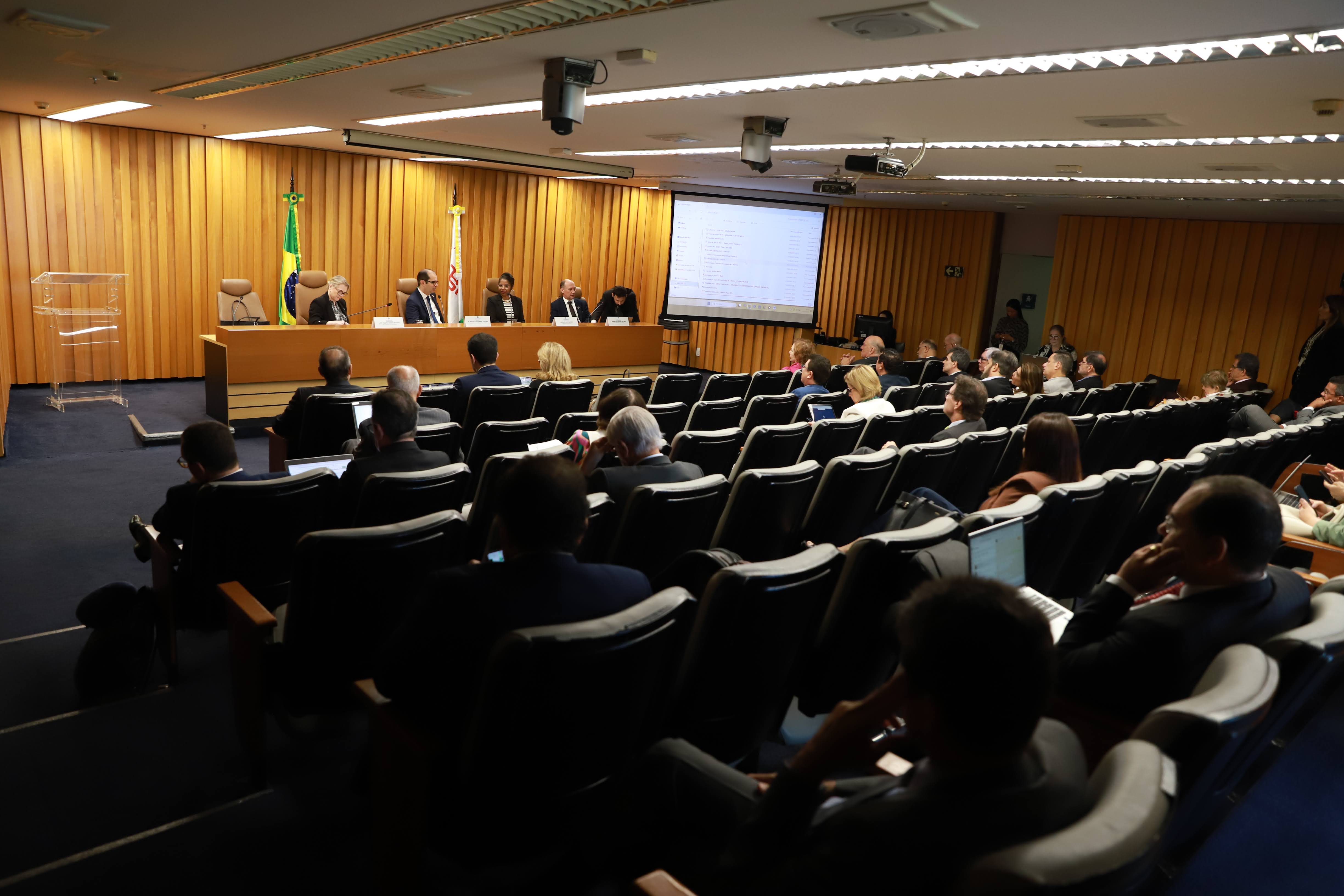 Foto de um auditório, com o público em primeiro plano e a mesa de trabalhos ao fundo. Aparecem ao fundo as bandeiras do Brasil e do Distrito Federal. Cinco pessoas estão na mesa. Na direita da imagem um telão está ligado.