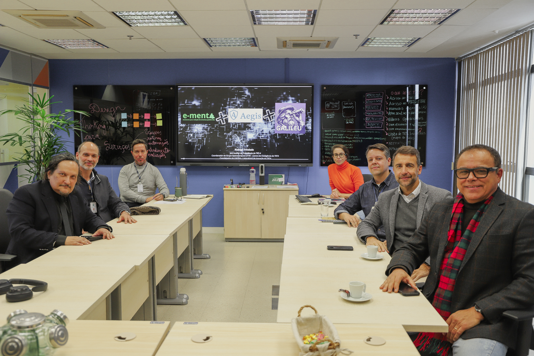 Grupo de sete pessoas, entre elas Gabriel e Rodrigo, reunidos em sala do Linova, com telas digitais e quadros ao fundo com as tecnologias desenvolvidas pelo Tribunal.