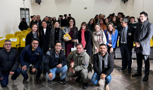 Um grupo de cerca de 25 pessoas posa sorrindo para a foto em um auditório. Ao centro, uma mulher segura uma cesta embrulhada com celofane. Há pessoas em pé ao fundo e outras agachadas à frente. O ambiente é iluminado e há cadeiras amarelas vazias atrás do grupo.