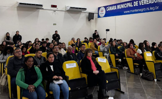 A imagem mostra o público sentado em cadeiras amarelas, assistindo ao evento na Câmara Municipal de Vereadores de Montenegro.