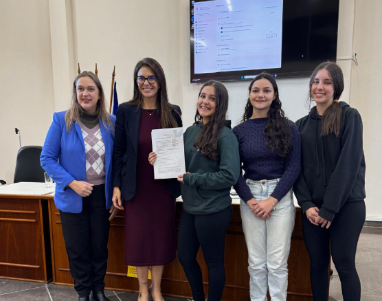 Cinco mulheres posam sorrindo. No centro, uma jovem segura um documento ao lado da juíza Aline Fagundes. Elas estão em frente a uma bancada de madeira, com bandeiras ao fundo e uma tela ligada acima delas. As jovens usam roupas casuais; a juíza e outra mulher vestem blazers.