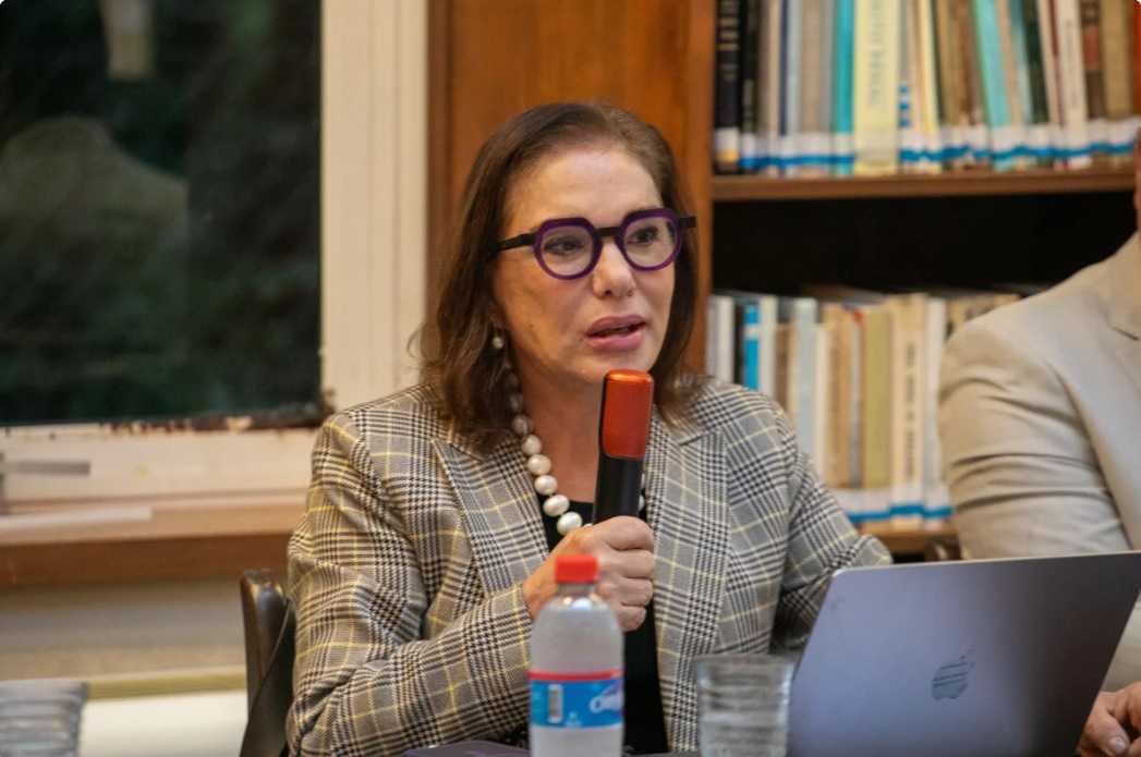 Judith Martins Costa é branca e tem cabelo castanho. Usa óculos de aro roxo, blazer creme xadrez e camisa preta. Está falando enquanto segura um microfone. Está sentada em uma cadeira e atrás dela aparece uma estante com livros.  0/500
Nenhum arquivo escolhido	>