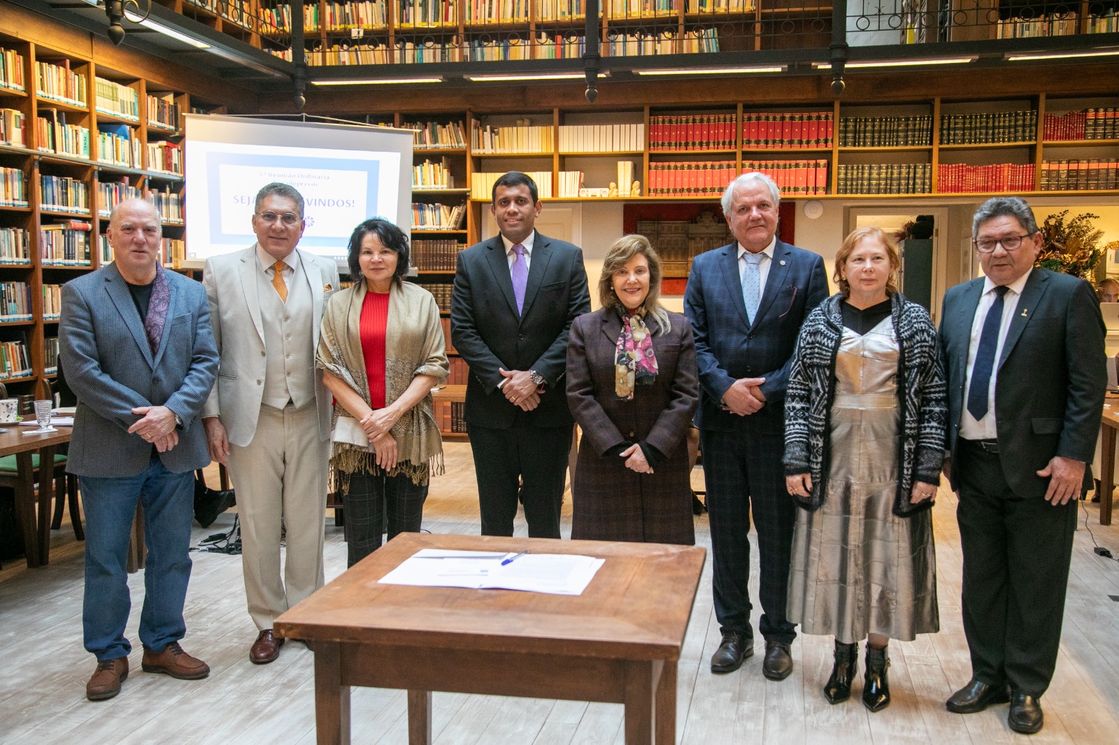Oito pessoas estão de pé sorrindo e posando para a foto. Na frente deles aparece uma mesa com um documento sobre ela e uma caneta. Ao fundo, as paredes são ocupadas por estantes altas repletas de livros.
