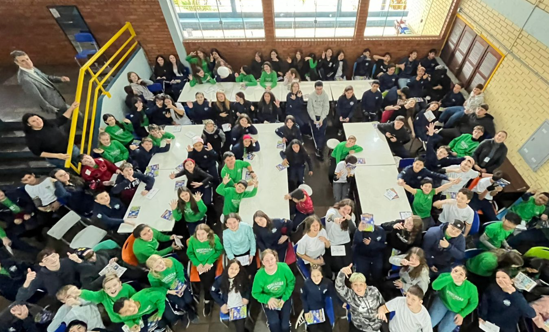 Uma grande quantidade de estudantes, muitos vestindo camisetas verdes e uniformes escolares, olha para cima e acena, sorrindo. Eles estão reunidos em um refeitório escolar, sentados em torno de várias mesas.
