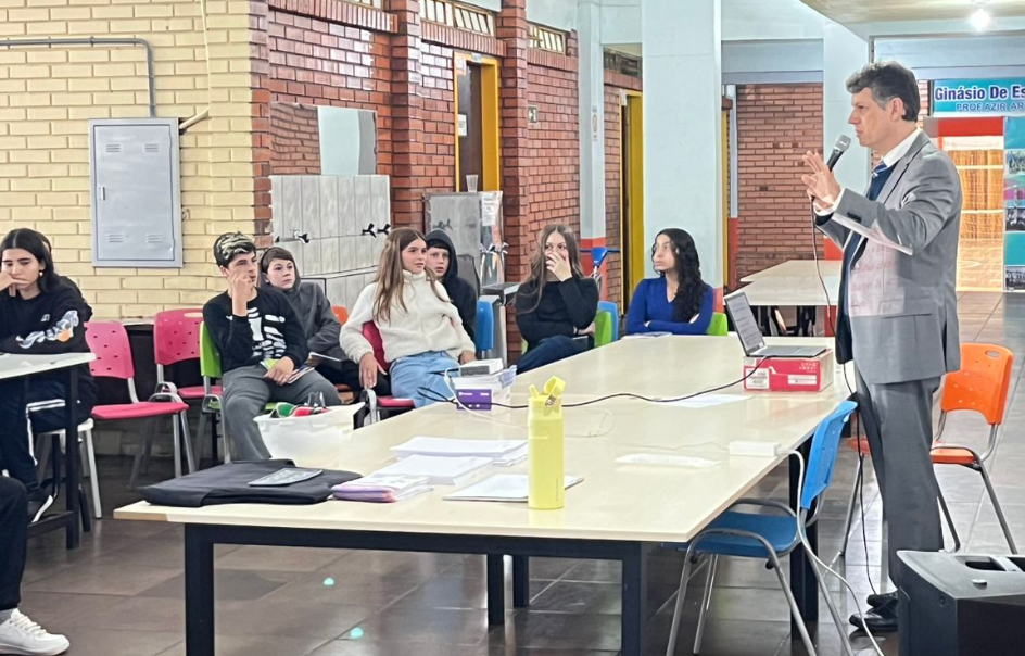 Juiz Marcelo está em pé, falando com microfone em mãos para um grupo de adolescentes sentados em volta de mesas em uma sala de escola.