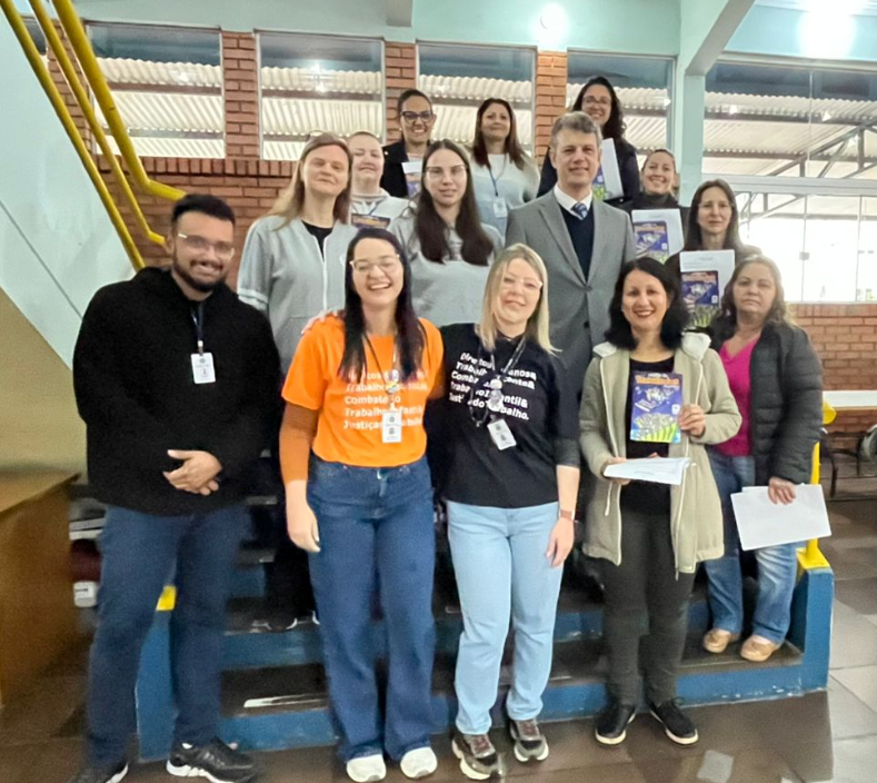 Um grupo de cerca de 15 pessoas posa para uma foto sorrindo em uma escadaria interna de uma escola. Na frente, algumas mulheres seguram cartilhas. O ambiente é iluminado e tem paredes de tijolos e escadas com corrimão amarelo.