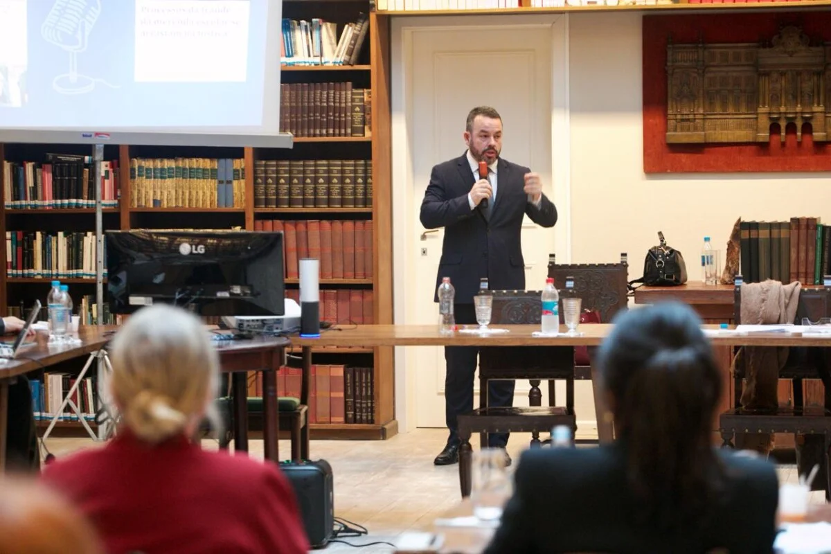 Eduardo Matos falando ao microfone em uma biblioteca , com estantes cheias de livros ao fundo. Ele está em pé, ao lado de uma mesa com garrafas de água e copos, e há um projetor exibindo slides. Na frente dele, algumas pessoas estão sentadas, assistindo à apresentação.