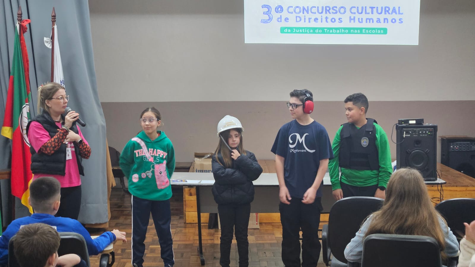 cinco estudantes em pé, alguns usando capacete, óculos e abafador. Ao fundo, há o telão com a identidade visual do "3º Concurso Cultural de Direitos Humanos da Justiça do Trabalho nas Escolas".