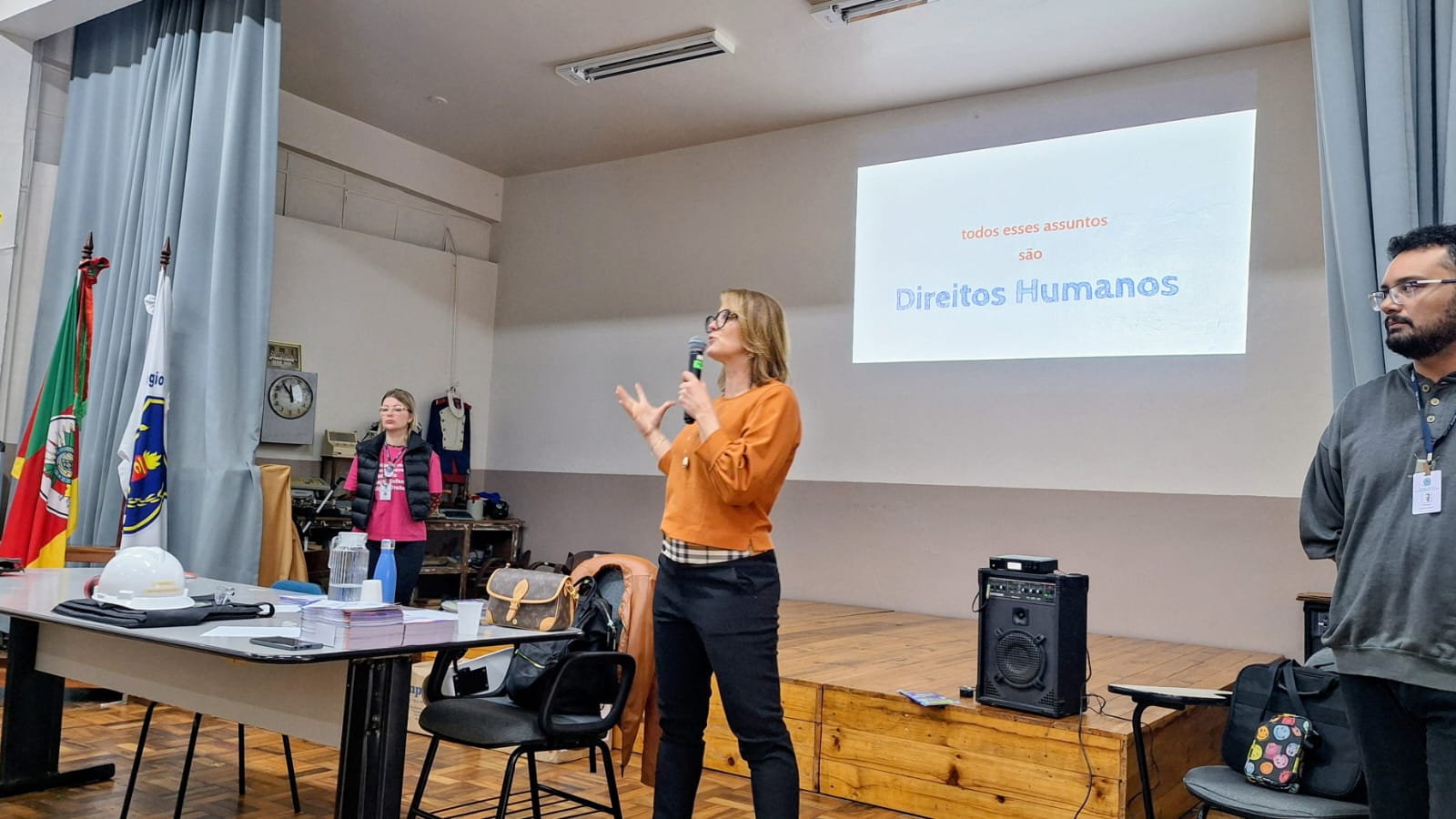 Juíza Adriana Kunrath fala ao microfone na frente de um telão com os dizeres "Todos esses assuntos são Direitos Humanos