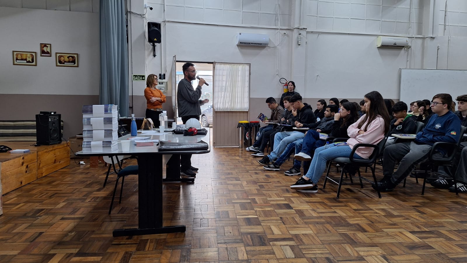 Juiz Marcelo posa ao lado de cinco estudantes que usam equipamentos de proteção como capacete, colete e abafador de ruído. Ao fundo, aparece um telão com a frase "Qualquer assunto relacionado aos direitos humanos no mundo do trabalho".