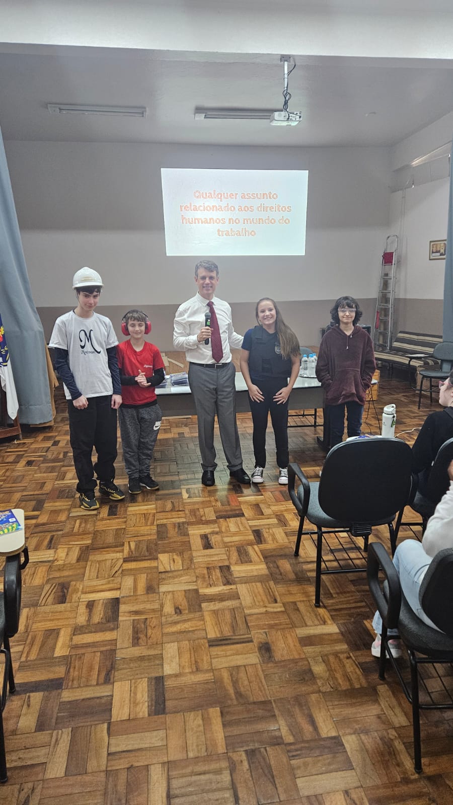 Um homem fala ao microfone em frente a uma turma de estudantes sentados. Ele está ao lado de uma mesa com materiais e cartilhas empilhadas. Ao fundo, uma mulher observa de pé, próxima à porta.