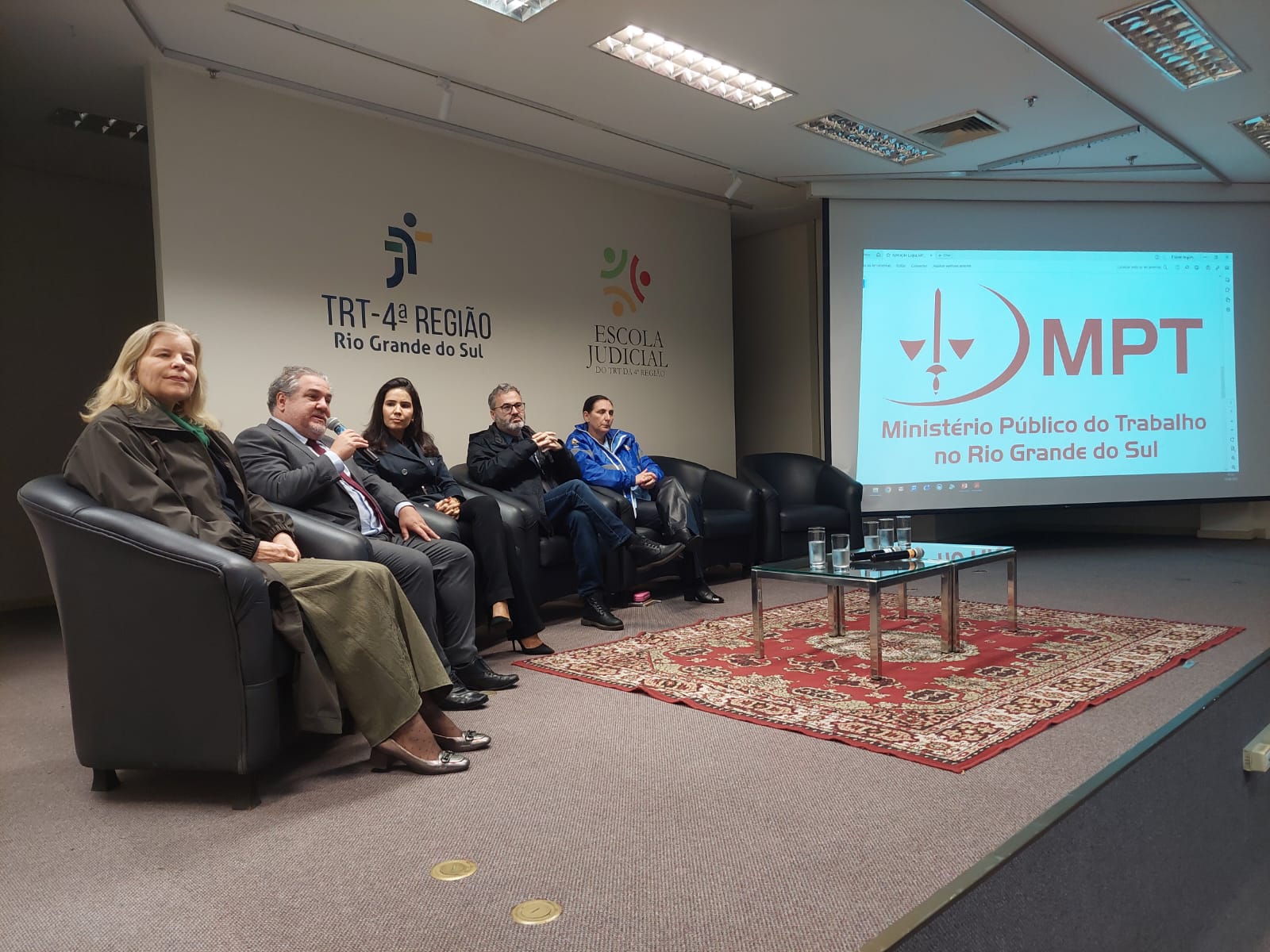 Um painel de cinco pessoas sentadas em cadeiras de couro pretas. Atrás delas, um telão projeta o logo do "MPT - Ministério Público do Trabalho no Rio Grande do Sul". Há um tapete e uma mesa de centro com copos de água na frente do painel. No fundo, há logos do TRT4 e da Ejud4.