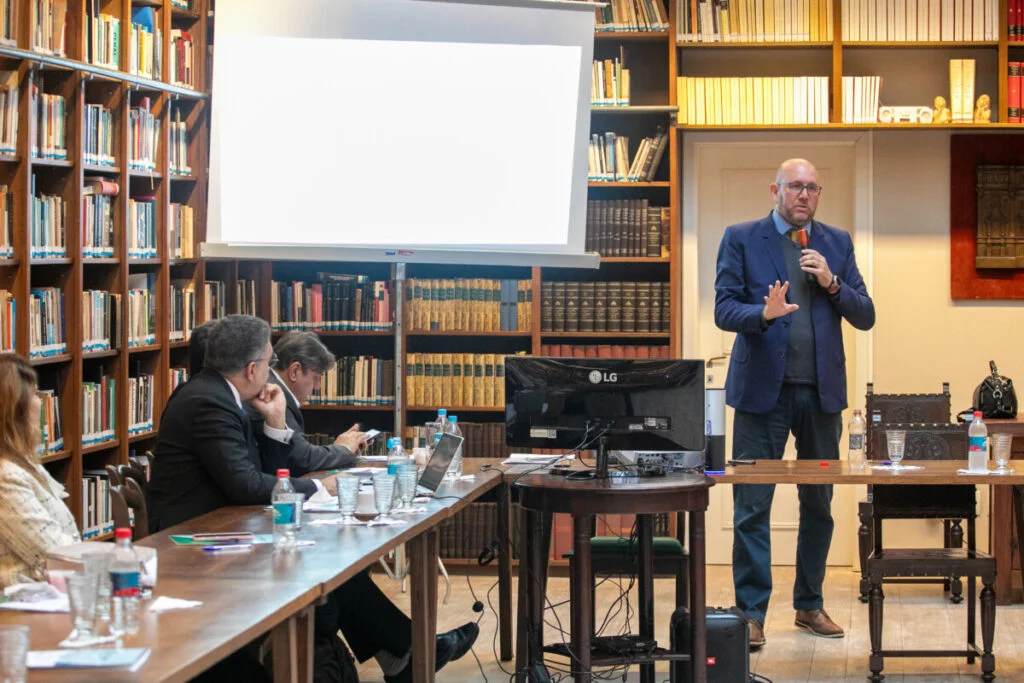 Servidor Marcelo Lucca em pé, segurando um microfone e fazendo uma apresentação em uma biblioteca, diante de uma mesa com outras pessoas sentadas e um projetor ligado ao fundo.