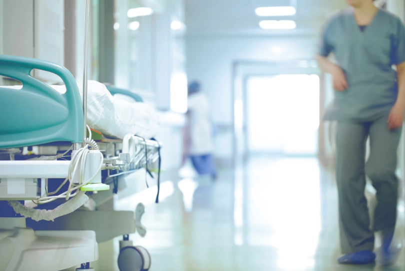 Mulher de pele clara e uniforme azul em corredor do hospital. Não há a imagem do rosto. Em primeiro plano, no canto da imagem, há uma cama hospitalar.