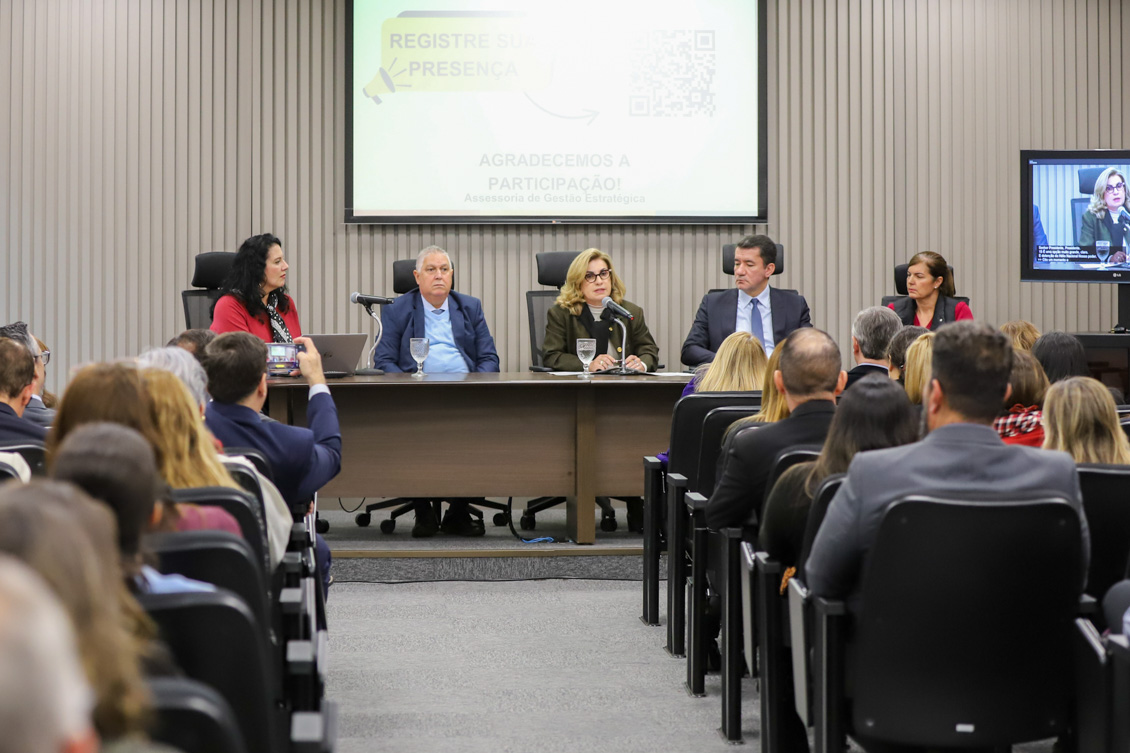 mesa de trabalhos composta pela Iara Gomes, Dr. Helcio, Dra. Ana Paula ao microfone, Dr. Renan e Dra. Regina. Todos sentados com o público em primeiro plano.