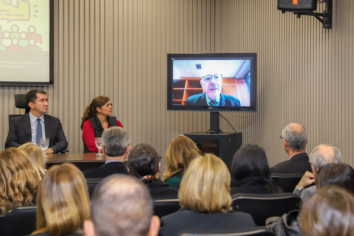 Participação do desembargador Paulo Mota em tela. Dr. Renan e Dra. Regina, sentados à mesa de trabalhos, observam a TV, bem como parte do público.