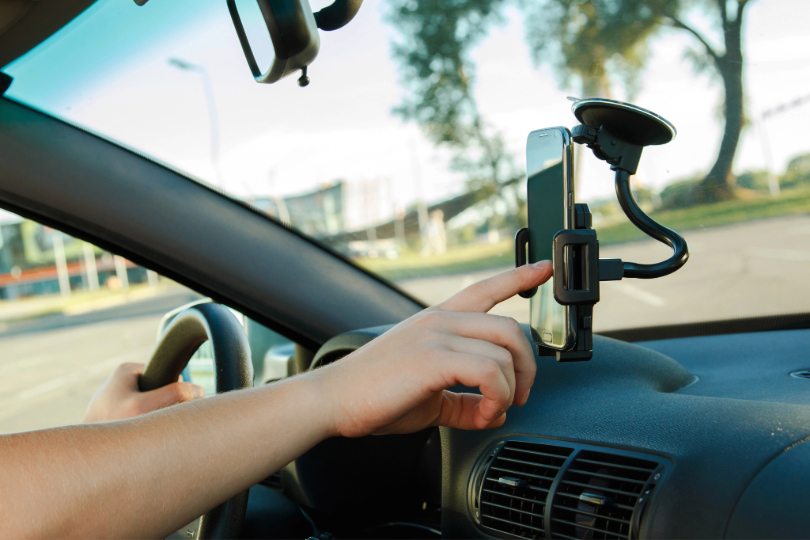 Mão masculina manuseia celular fixado ao para-brisa de um carro.