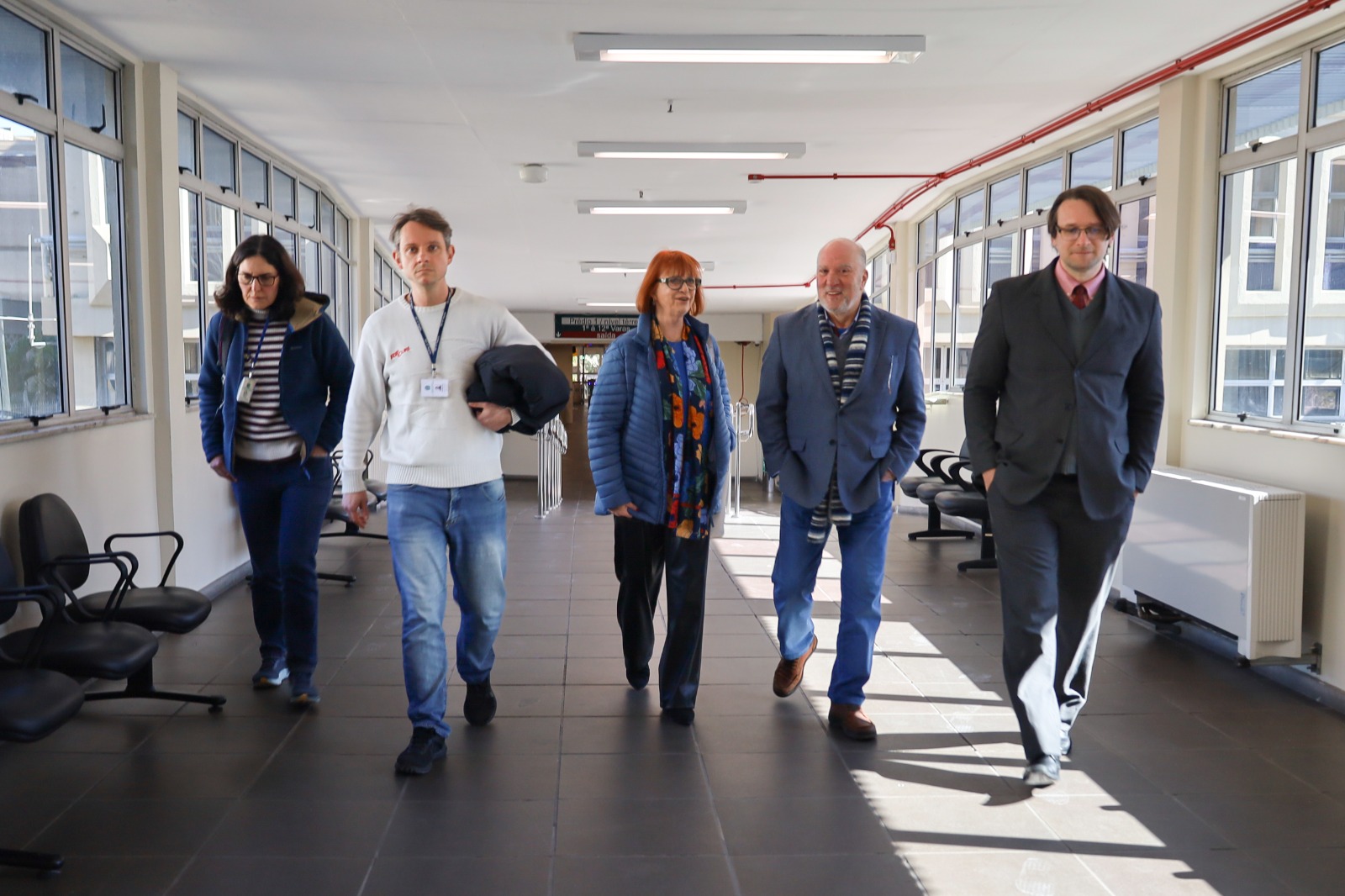 Cinco pessoas aparecem caminhando em uma galeria. Nas laterais aparecem janelas abertas. Elas andam em direção à câmera, sorrindo e conversando.