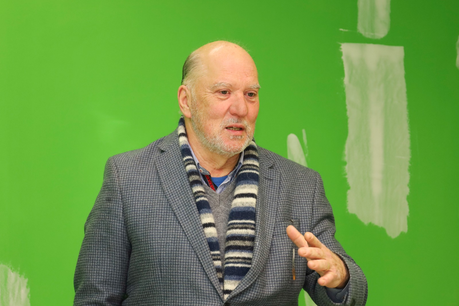 Martis Costa é branco, calvo e grisalho. Está usando um blazer cinza e uma manta. Fala e gesticula. Atrás aparece uma parede verde usada como cromaqui. 