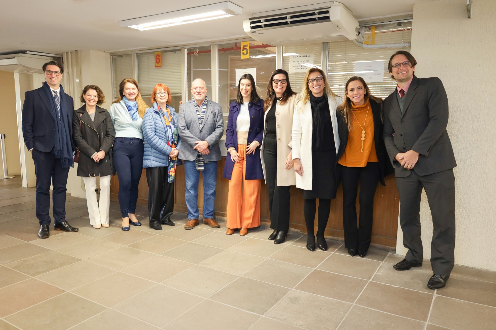 Dez pessoas posam para a foto. Elas estão de pé, sorrindo, em frente a um guichê de atendimento que está fechado. 