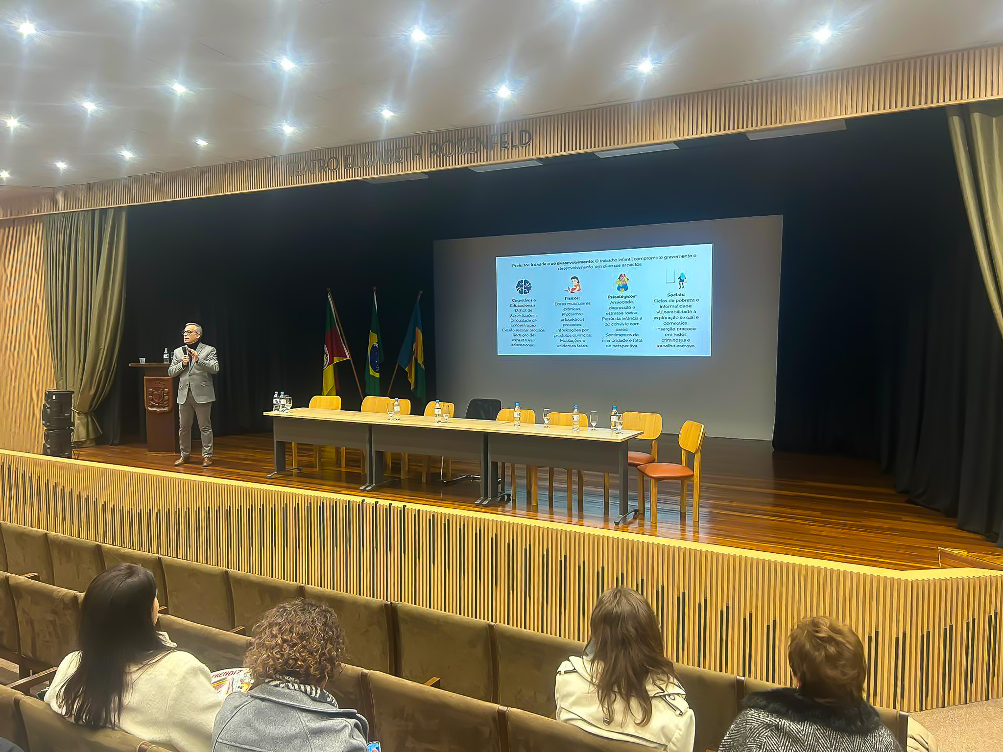 Palestrante apresenta dados sobre os impactos do trabalho infantil. Slide projetado ao fundo, com público assistindo.
