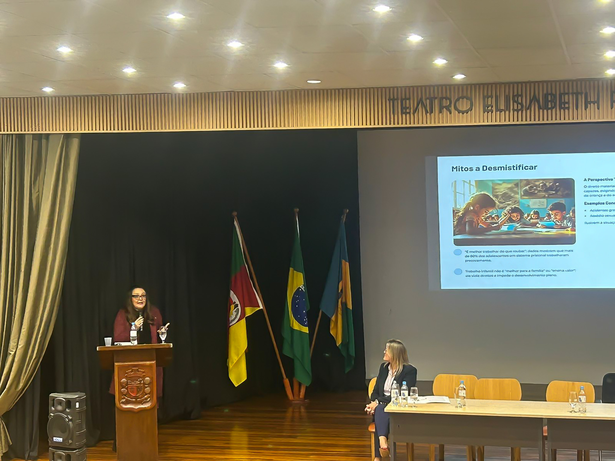 Apresentação sobre mitos do trabalho infantil no Teatro Elisabeth Rosenfeld. Mulher fala ao púlpito, enquanto outra pessoa assiste sentada. Slide projetado ao fundo e bandeiras ao lado.