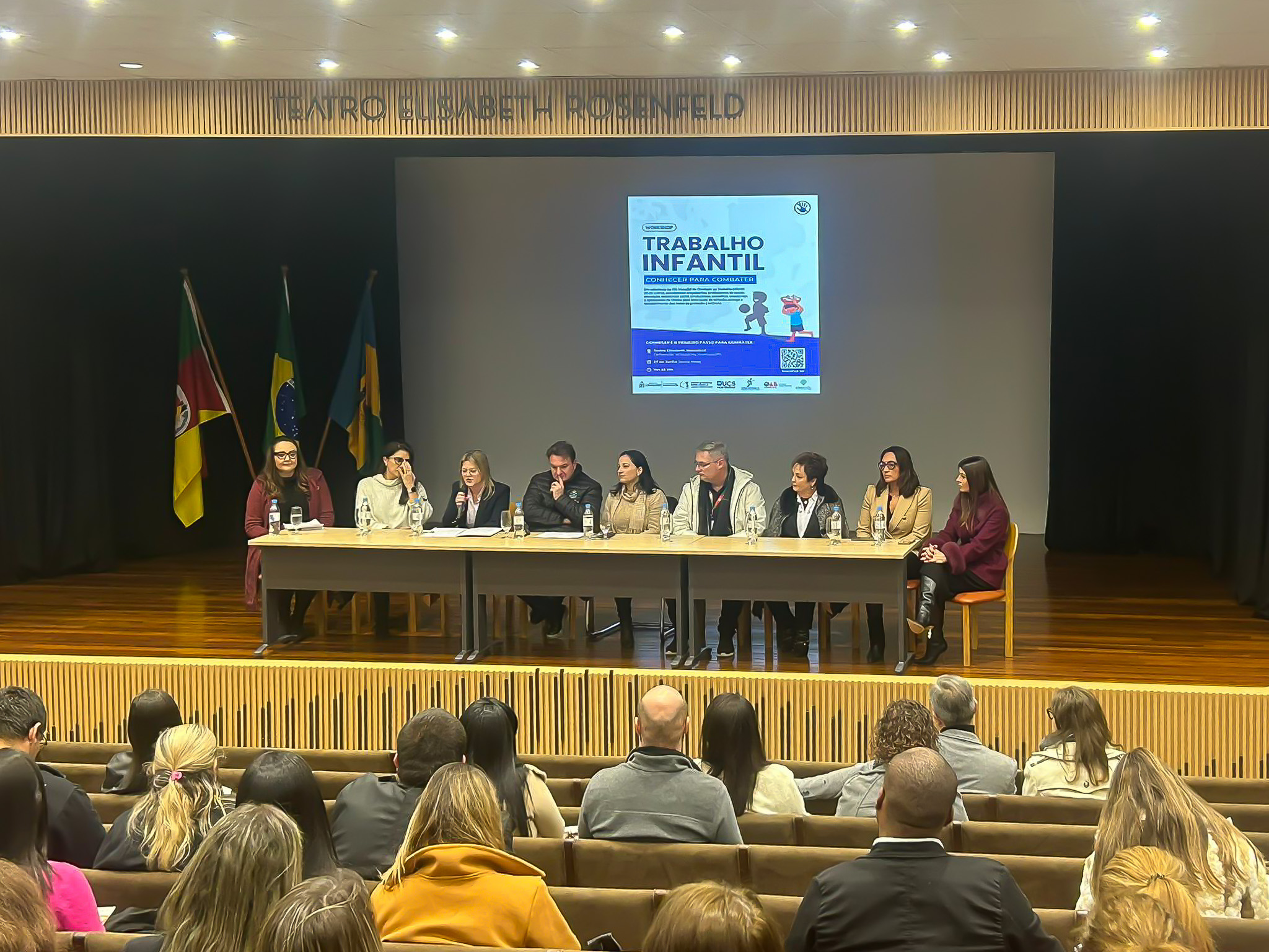Evento sobre trabalho infantil no Teatro Elisabeth Rosenfeld, com 11 palestrantes em mesa no palco e público assistindo. Cartaz do evento ao fundo e bandeiras expostas.