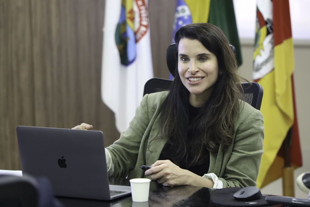 Carolina é branca e tem cabelo castanho liso e comprido. Veste blusa preta e casaco verde. Atrás dela aparecem as bandeiras de Poa, do Brasil e do RS. Ela fala e gesticula, sorrindo, em frente a um notebook aberto. 