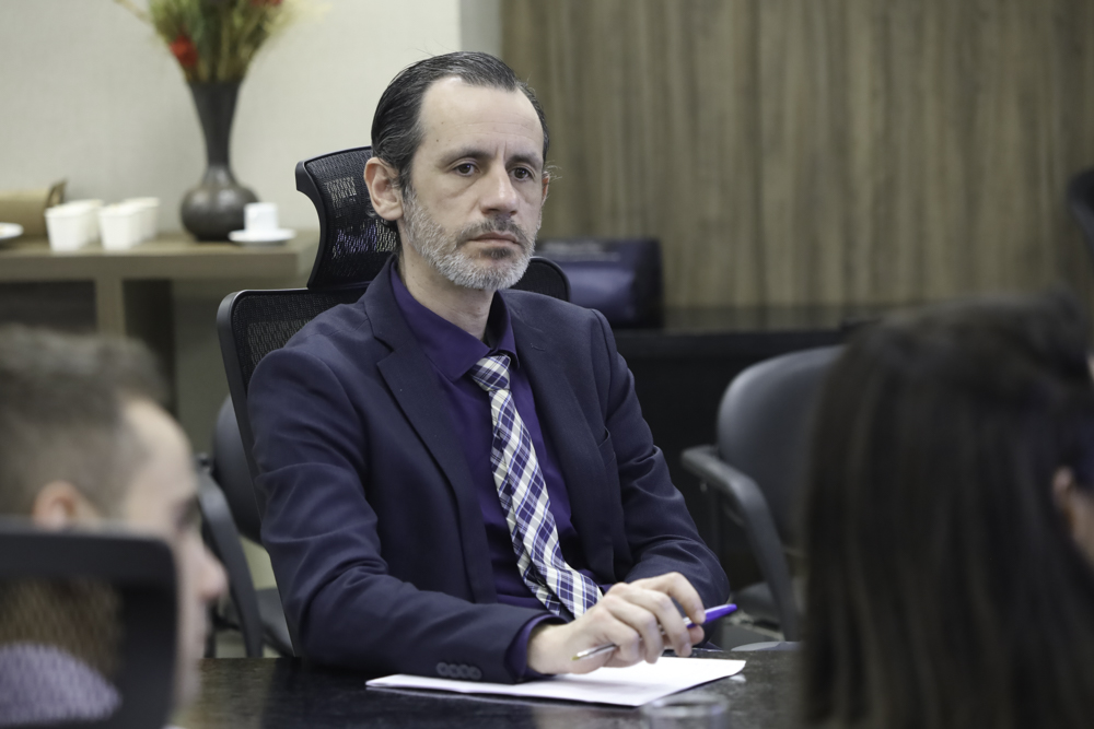 Rafael Brandolf é branco, tem cabelo escuro e barba grisalha Usa terno e camisa escuras e gravata xadrez. Está sentado olhando para o lado direito, prestando atenção em quem fala. 