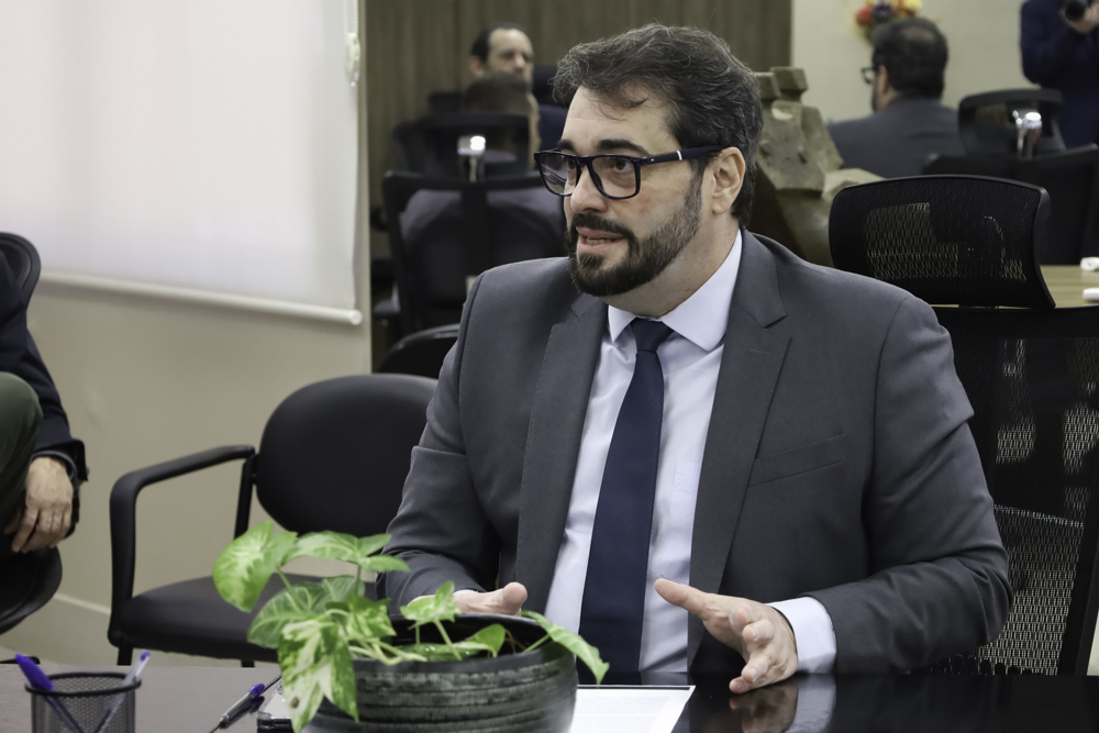 Alexandre é branco, tem cabelo e barba escura. Está sentado falando e gesticulando. Usa terno cinza, camisa branca e gravata azul. 