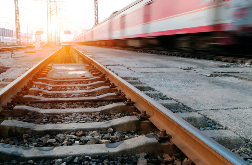 linha férrea com trem se aproximando. Ao lado, um trem vermelho e cinza em alta velocidade.