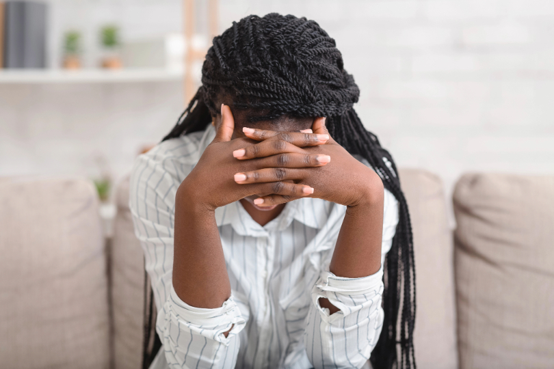 Mulher negra tapa o rosto com as mãos, demonstrando tristeza. Ela tem longos cabelos trançados e usa camisa branca e calça jeans