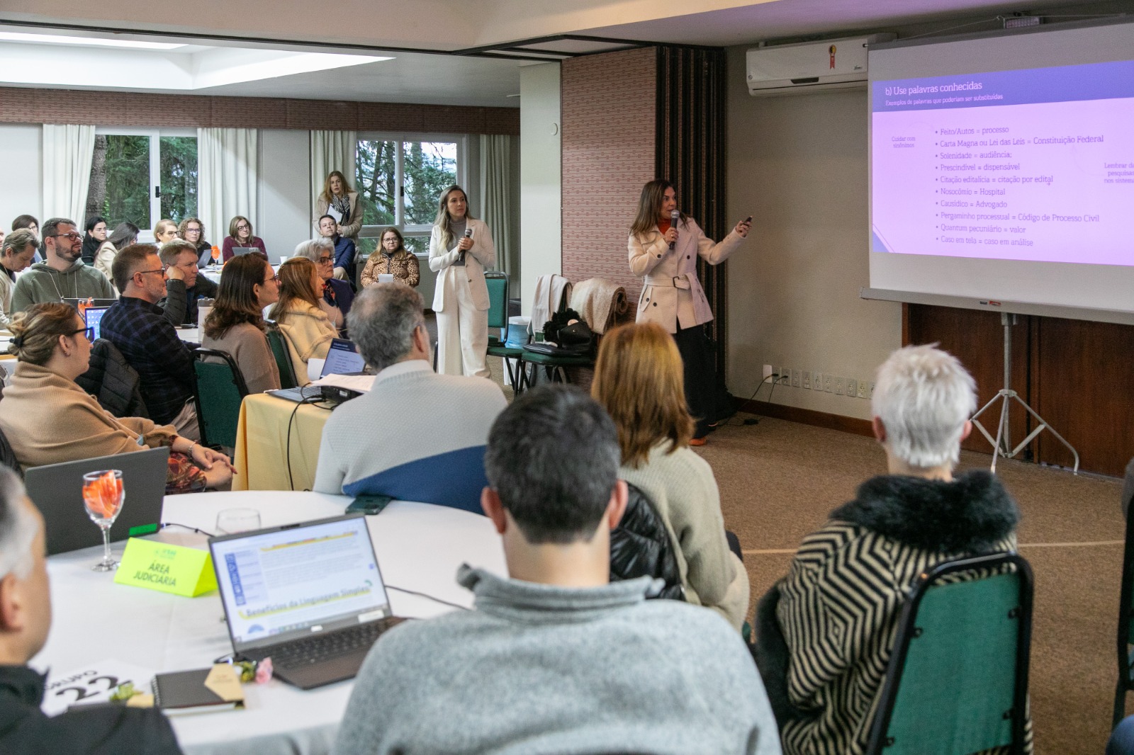 Público em primeiro plano, sentados em mesas e olhando a apresentação. Uma mulher aparece de pé falando e apontando para um telão.
