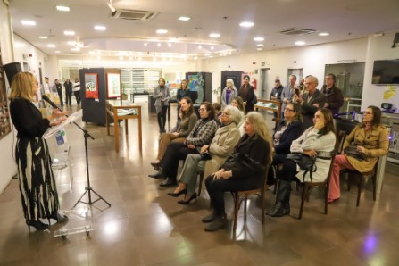 Marilia Fayh em pé falando ao microfone diante de convidados sentados em cadeiras no saguão do TRT4