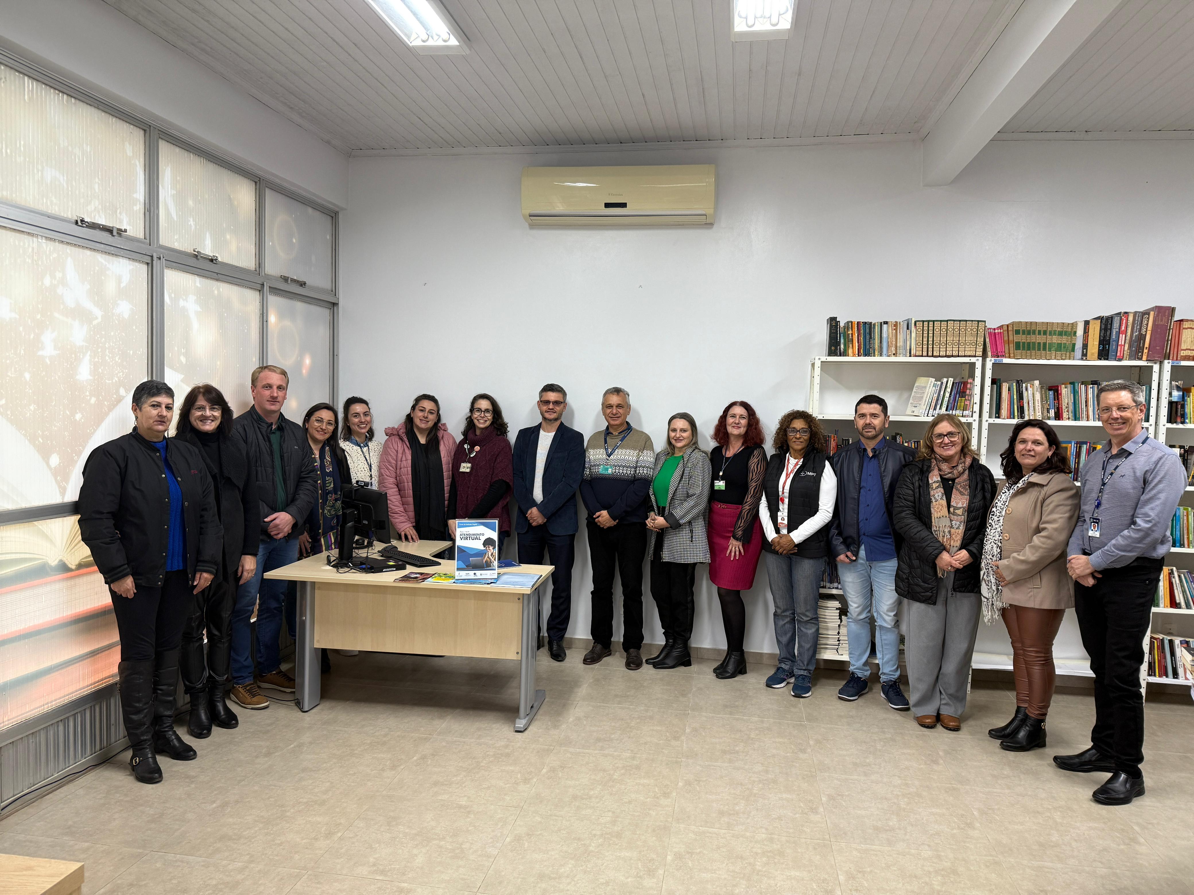 Grupo de 18 pessoas posa em visita institucional a uma biblioteca, com estantes de livros ao fundo e mesa com material informativo em destaque.