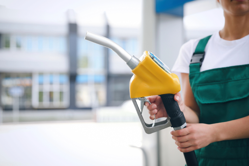 Jovem frentista segura bico de bomba de gasolina. Ela usa macacão verde e camiseta branca. Não há imagem do rosto