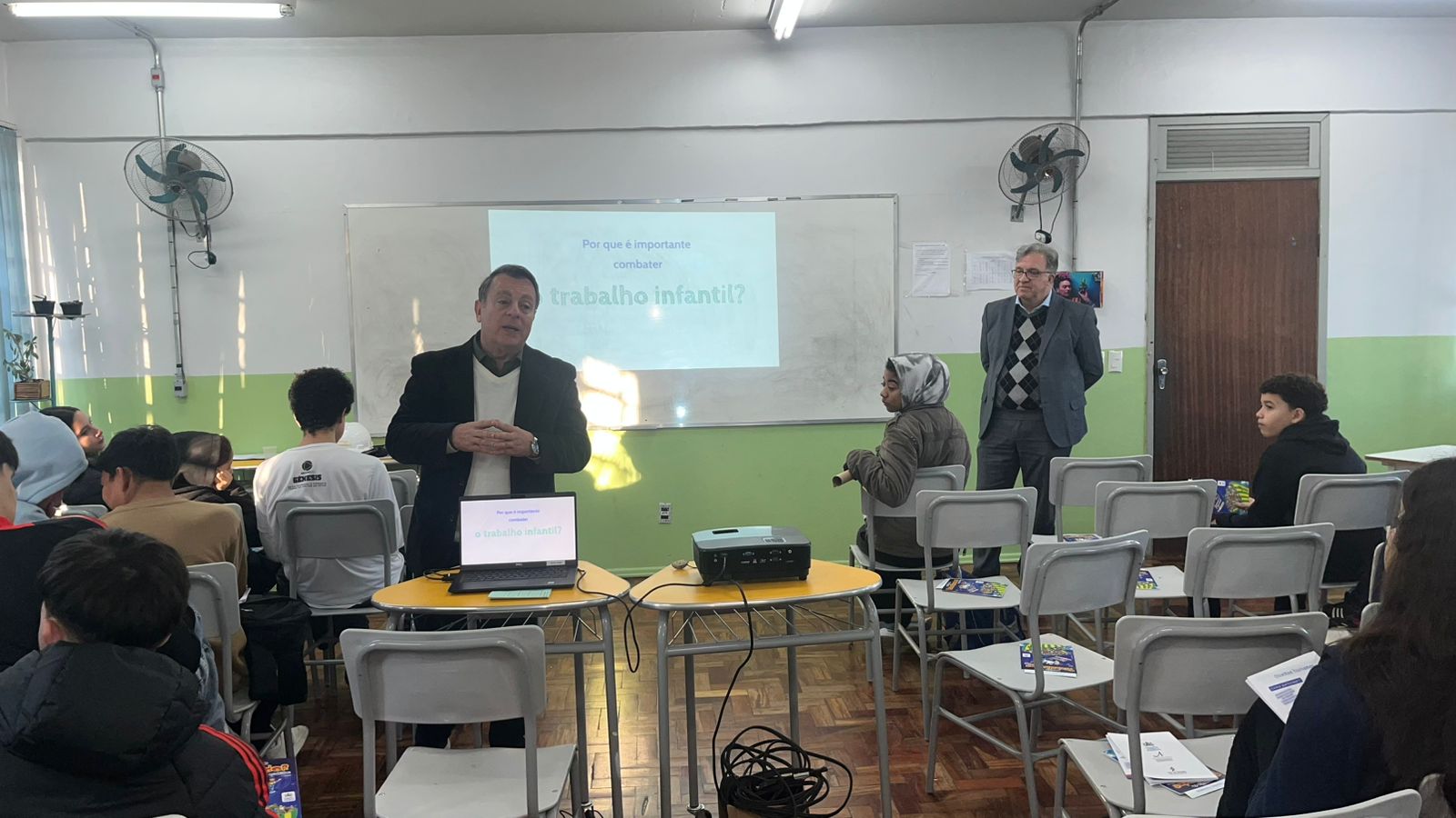 Sala de aula com alunos sentados de costas assistindo a uma apresentação. Ao fundo, no quadro, está projetada a pergunta: “Por que é importante combater o trabalho infantil?”. Márcio e Cassou falam com os estudantes; um deles está em pé ao centro e o outro próximo à porta. Sobre as mesas, há um notebook e um projetor.