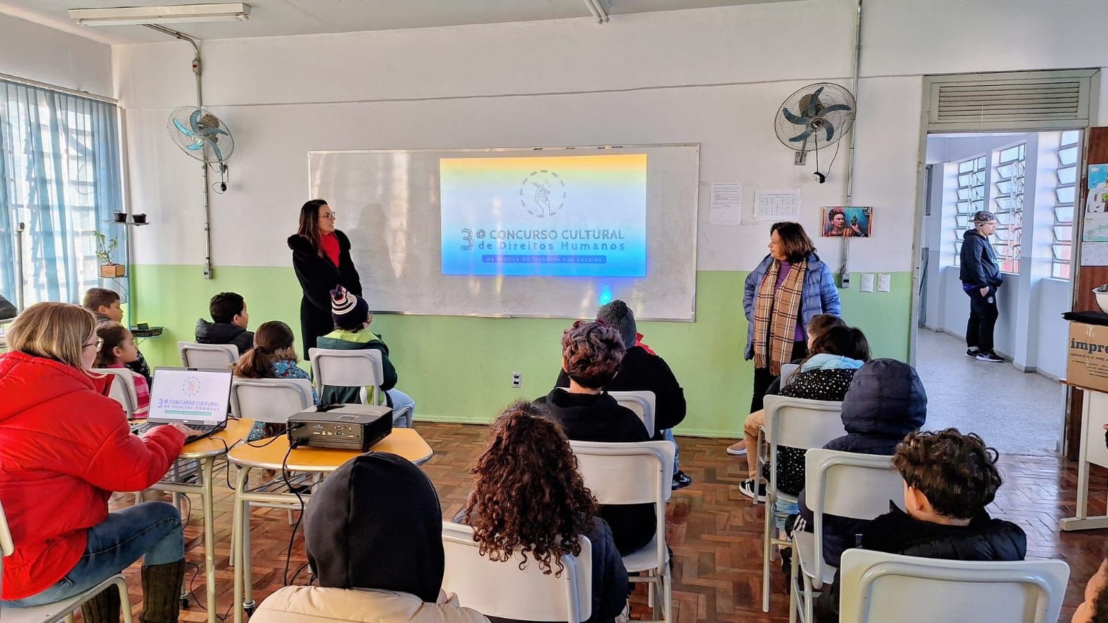 Alunos sentados em carteiras escolares assistem a apresentação sobre o 3º Concurso Cultural de Direitos Humanos. A logo do concurso é projetada no quadro branco. Carolina e Rejane estão de pé conversando com os estudantes.