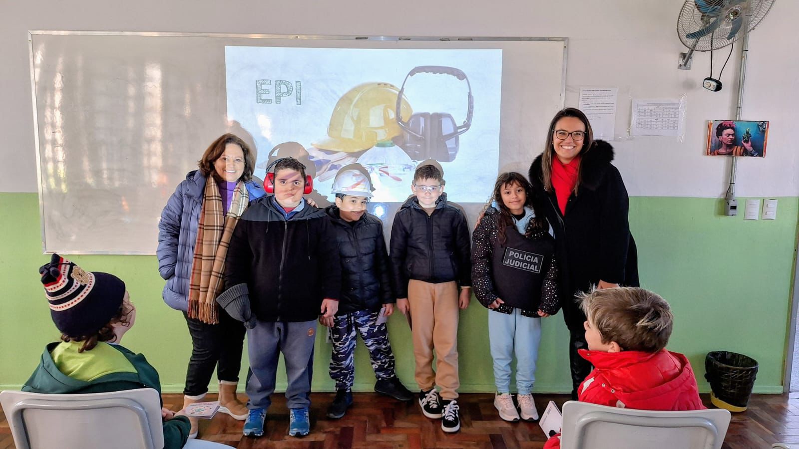 Quatro crianças, Rejane e Carolina posam para foto em frente a uma projeção com o tema “EPI”. As crianças usam equipamentos como protetor auricular, capacete, óculos de proteção e coletes. Uma das crianças veste um colete escrito “Polícia Judicial”.