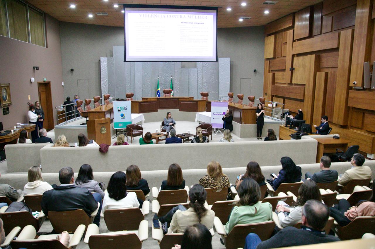 Vista geral do plenário do TRE-RS durante evento sobre violência contra a mulher. Uma palestrante está sentada ao centro, com intérpretes de Libras ao lado e público assistindo nas cadeiras.