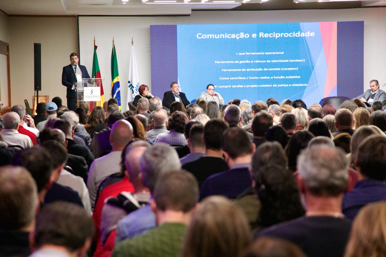 Público em primeiro plano. ao fundo, Diogo fala no púlpito, ao lado de bandeiras do Brasil, RS e TRT-RS. No palco aparecem, sentados em poltronas, Bárbara, Aldo, Rejane e Adolfo.