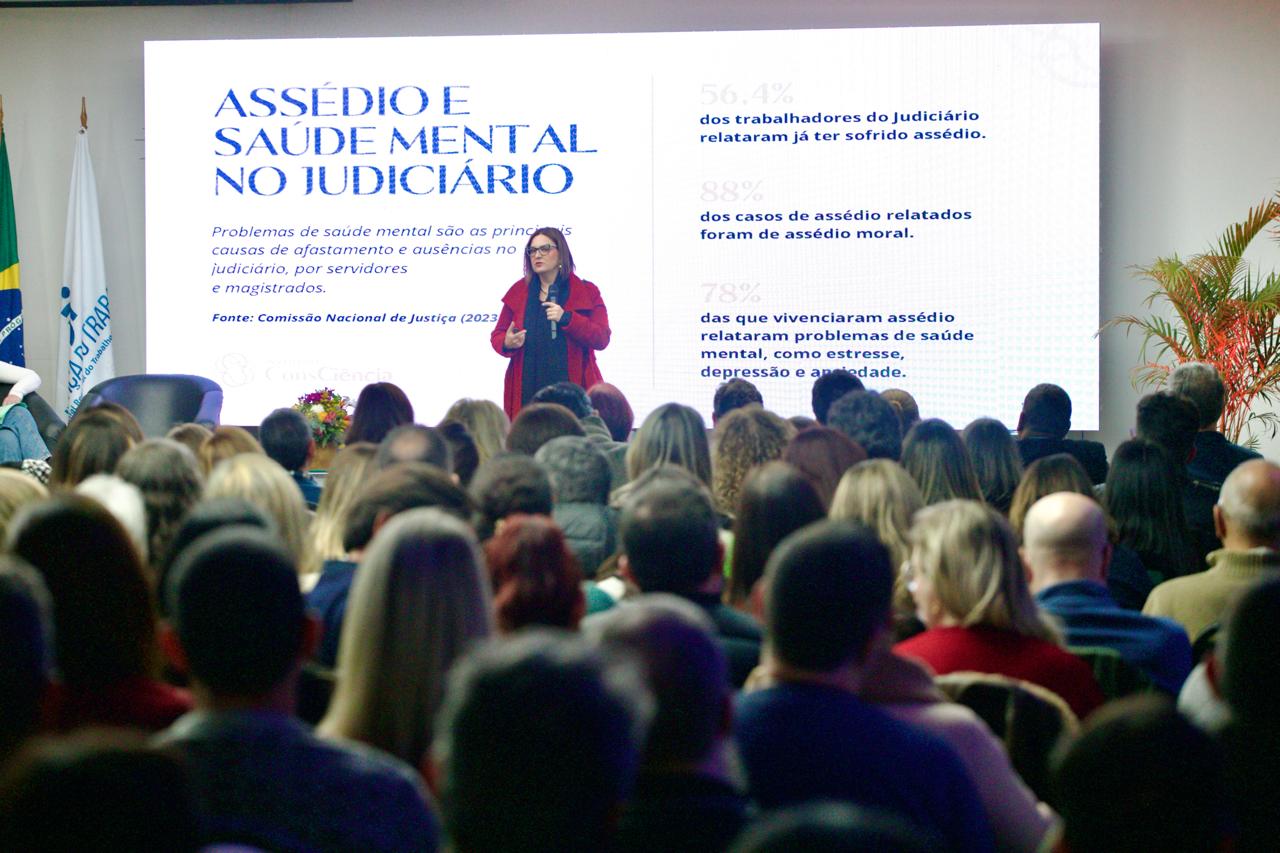 A foto mostra a palestrante Lis ao fundo, com público na frente e o telão aparecendo a frase assédio e saúde mental no Judiciário.