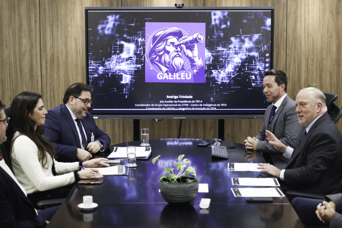 Quatro pessoas conversam em uma mesa de reuniões, com um telão mostrando o logo do Galileu ao fundo. O logo mostra uma pessoa de barba usando uma luneta.