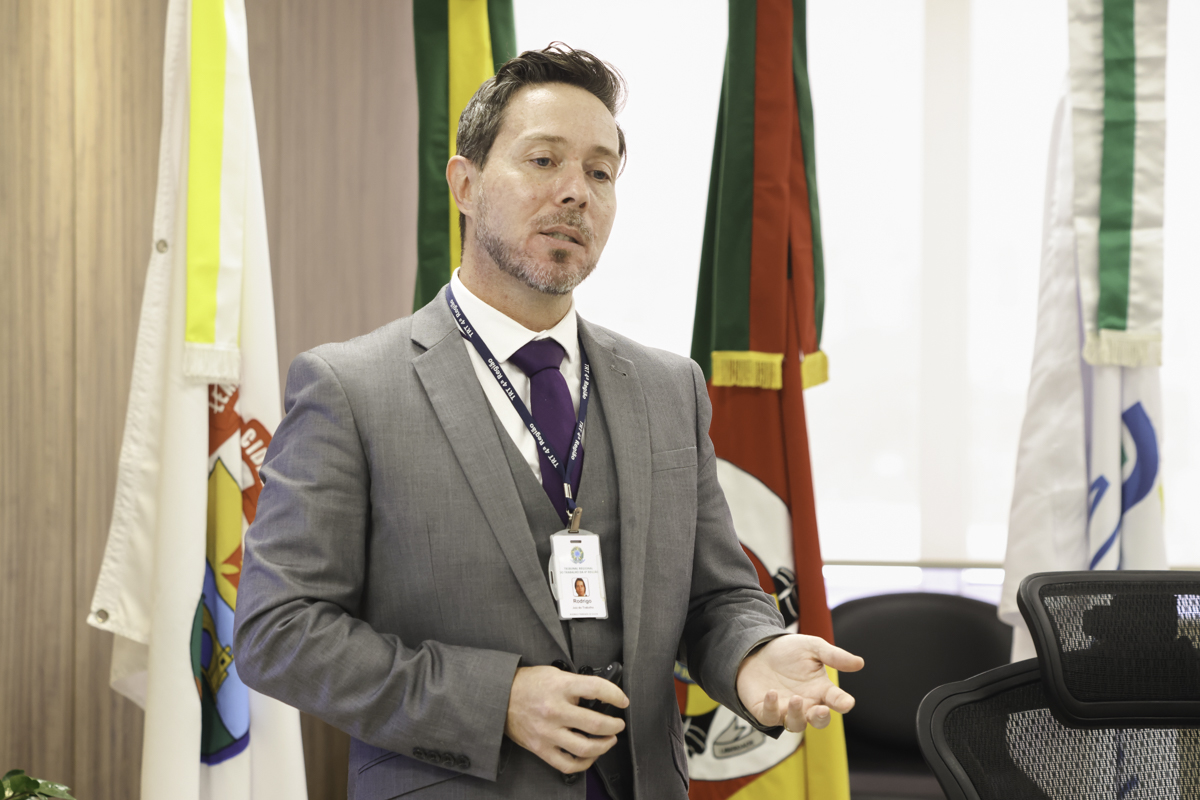 Rodrigo é branco, usa cabelo castanho curto e barba curta. Veste terno com colete cinza, e gravata bordô. Fala e gesticular. Ao fundo aparecem as bandeiras de Poa, do Brasil, do RS e da JT.