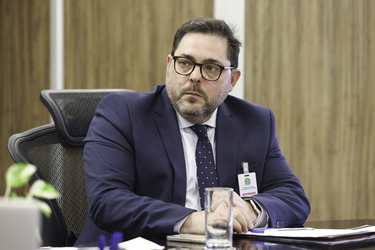 Camilo é branco, de cabelo castanho curto  e usa óculos de aro escuro. Veste terno azul marinho e gravata azul com detalhes em branco e vermelho.