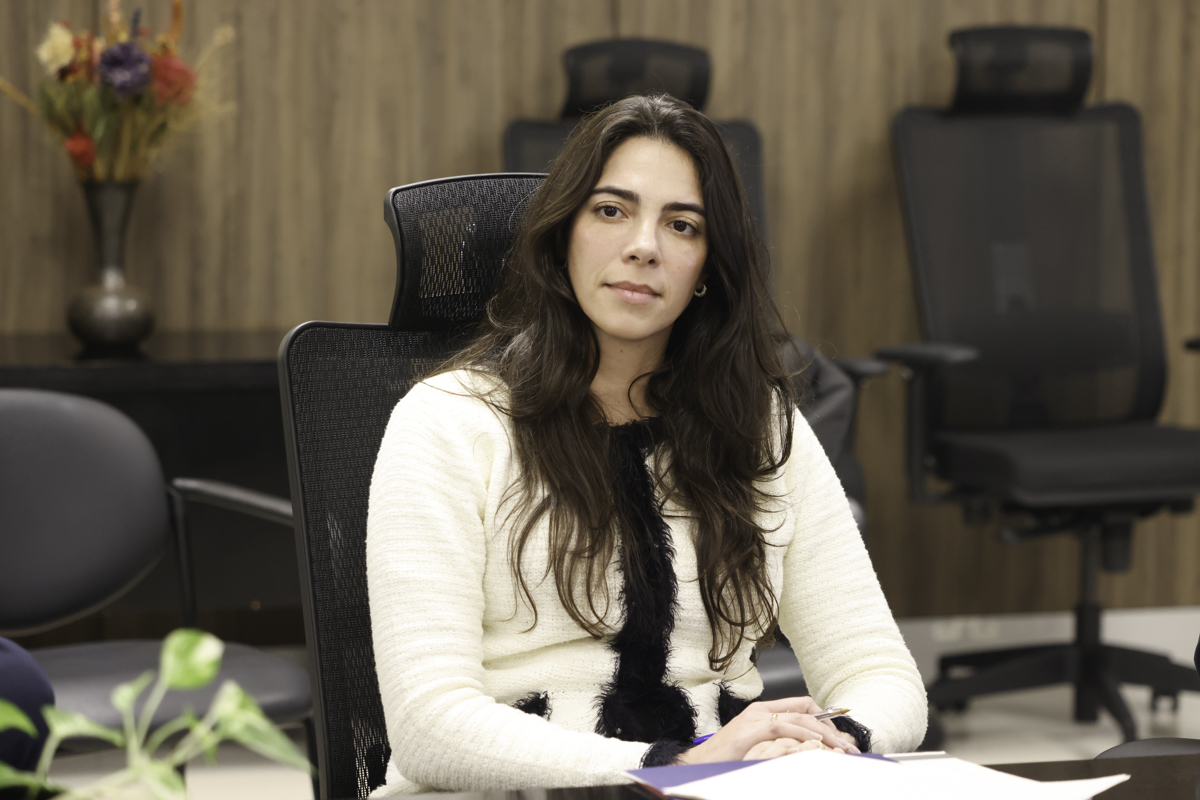 Mariana é branca, usa cabelo escuro liso e comprido. Veste uma blusa branca com detalhes em preto. Está com as mãos descansadas sobre a mesa, olhando para a esquerda da imagem.