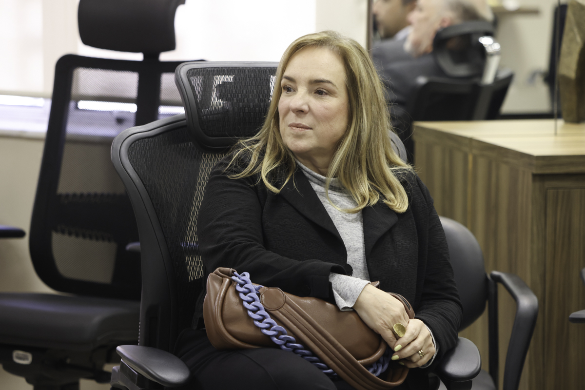 Maria Silvana é branca e loira. Usa bluza cinza e casaco preto. Está sentada, olhando para a esquerda da imagem e segurando uma bolsa marrom.