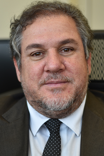 Salomão é branco, de cabelo e barba grisalha. Usa blazer escuro, camisa branca e gravata azul com pontos brancos. Está sorrindo para a foto.