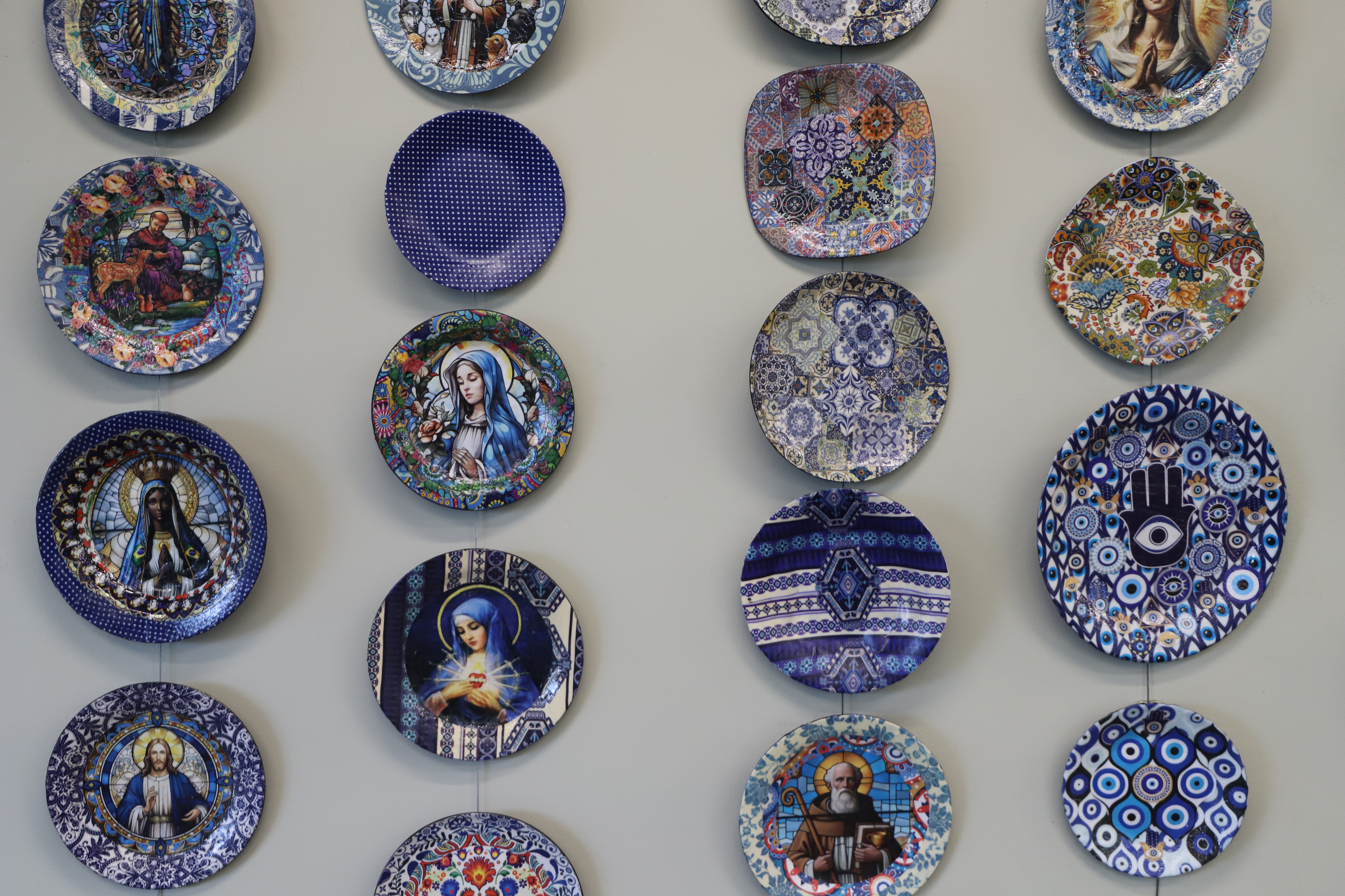 Pratos pendurados na parede, com imagens sagradas, figuras florais e geométricas. Os pratos são redondos, ovais ou quadrados, com fundo predominantemente azul.