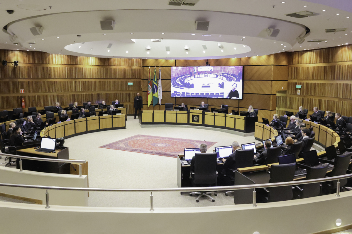 Nave do Plenário. Os desembargadores aparecem sentados em suas bancadas, que formam um grande círculo. O presidente está ao fundo, no centro da imagem, e na frente de sua bancada aparece o brasão da república. Acima dele há um telão exibindo a transmissão da sessão.